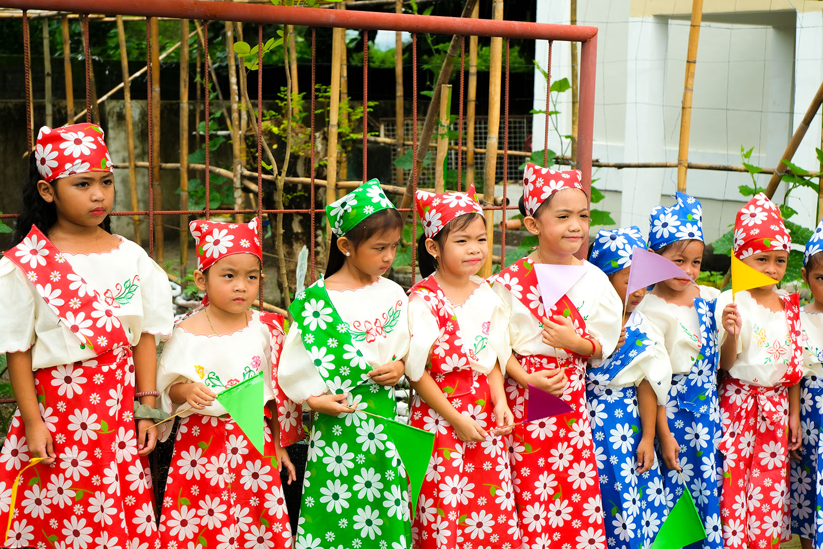 Girls in red, green, and blue floral Filipiniana costumes holding paper pennants at San Francisco Elementary