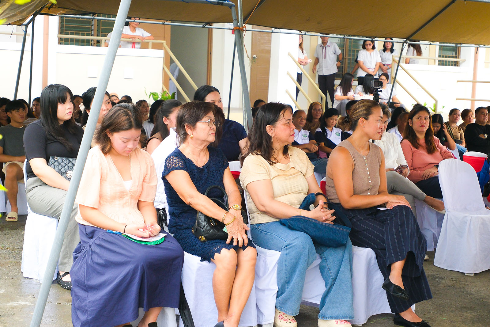 Parents and community members seated under the tent as a videographer films the ceremony